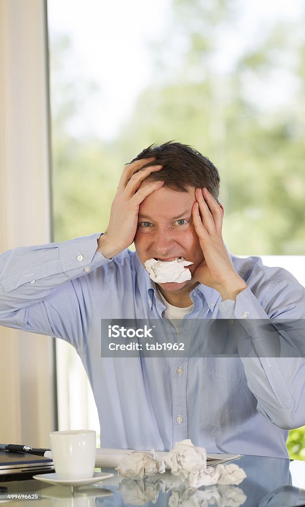 Mature man expressing rage at work Vertical image of mature man showing stress by biting wad of paper while working from home with bright daylight coming in from window in background Adult Stock Photo