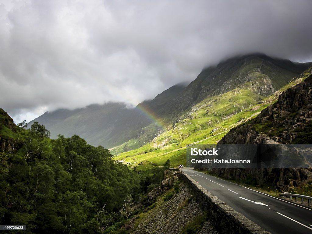 Road to independence Road in beautiful scottish mountains with heavy clouds and rainbow above it Beauty Stock Photo