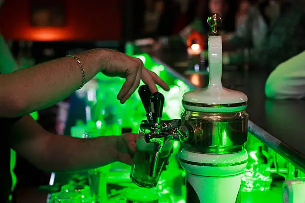 Bar in the nightclub. Beer podium. In a glass of beer is poured. Blurred outlines of visitors. Nightlife in the kluyuche. Green backlit locker with alcohol.