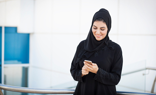 Young arabian female entrepreneur is standing outside white office building and using her smartphone to read e-mails, chat with friends, surf the net and manage business communication on internet platforms. She is smiling and looking at phone screen. Wearing traditional arabic clothes for women. Image contains copy space. Made in Dubai, UAE.