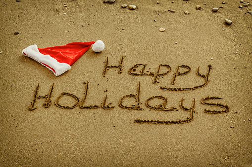The words Happy Holidays written in wet sand with a Santa Hat on a beach