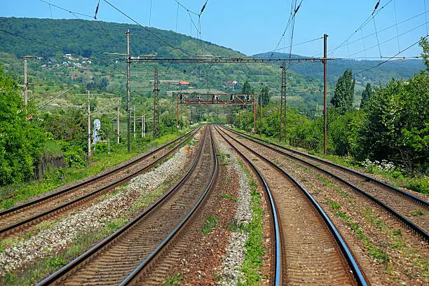 Four mainline tracks on an electrified railway pass under a signal gantry with one red and one green light then head into the distance and round a corner.  In the background, a line of hills.  Taken from the rear of a train.  No trains in the image.  Horizontal, copy space.