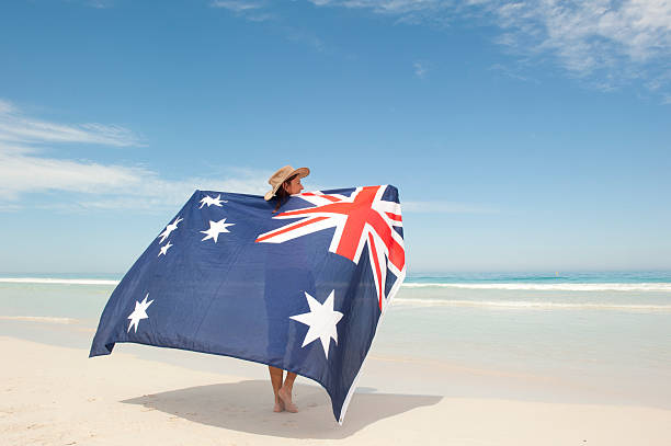 Attractive mature woman wearing akubra hat and with Australian flag around shoulder standing at tropical Australian beach, isolated with ocean and blue sky as background and copy space.