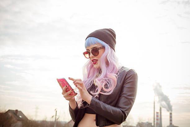 Outdoor portrait of blue-pink hair cool girl texting on phone Outdoor portrait of blue-pink hair cool girl wearing black leather jacket, beanie and sunglasses, texting on smart phone. Industrial zone in the background. cool attitude stock pictures, royalty-free photos & images