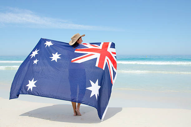 Attractive mature woman wearing akubra hat and with Australian flag around shoulder standing at tropical Australian beach, isolated with ocean and blue sky as background and copy space.