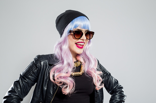 Portrait of excited blue-pink hair young woman wearing black leather jacket, beanie and sunglasses. Studio shot, one person.