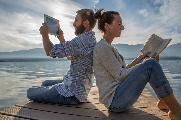 istock Couple sits on jetty above lake, uses book/digital tablet 499747952