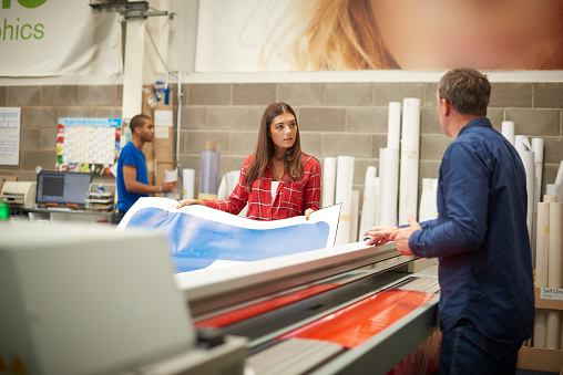 A young woman is working on a digital printing machine . her boss is explaining the machine or the job that she is doing . In the background rolls of vinyl can be seen , and a young co-worker is cutting out some vinyl lettering .