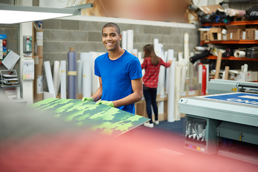 A young man is working on a digital printing machine and  handling the print for inspection .  In the background rolls of vinyl can be seen , and a young co-worker is choosing some vinyl .