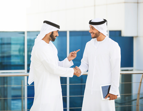 Couple of young arab men are meeting outside financial building. One of them is holding a digital tablet in his hand. They are shaking hands and gesturing, smiling. Both are dressed in traditional arabic style clothing for men. Blue glass windows of the building are in the background. Image was made in Dubai, UAE.