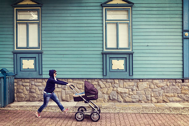 Woman in hoodie with baby stroller on walk against an old cyan house. She has just left the house. Spring time, Krynica Zdroj, Poland. Processed for retro look.