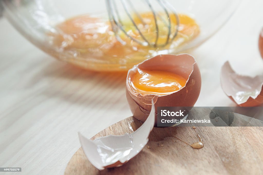 Cracked egg atop wooden kitchen counter. Close up of raw egg with glass bowl and a whisk in the background. Animal Egg Stock Photo