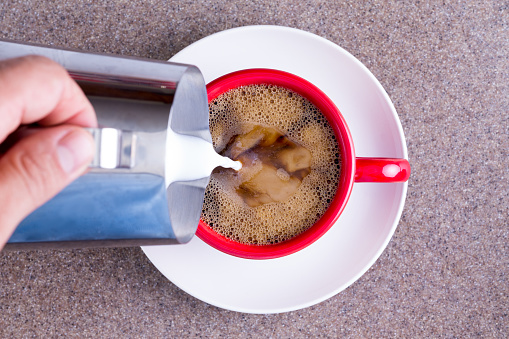 Man pouring milk from a jug into a cup of freshly brewed black filter or espresso coffee in a red mug on a white saucer against a grey background, overhead view