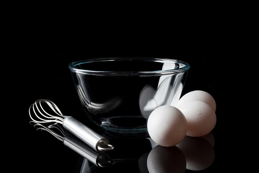 Glass transparent bowl with metal whisker and three white eggs aside from side on black background