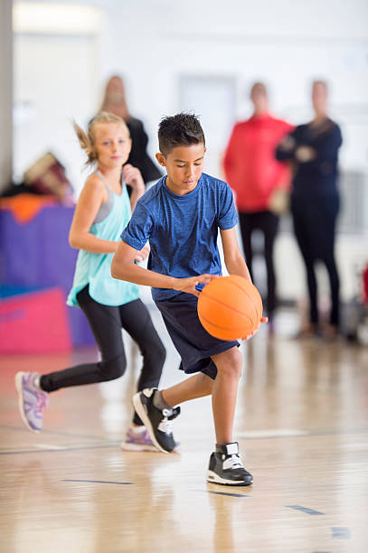 A multi-ethnic group of elementary age children are playing basketball together in the gym. They are dribbling the ball up the court.