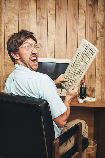 A man and office in 1980's - 1990's style, complete with vintage computer and technology of the time, yells in anger and frustration at his broken computer, ready to smash the monitor with his keyboard.  Wood paneling on the wall in the background.  Vertical image with copy space.