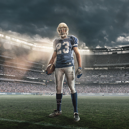 A low angle dramatic portrait of an American football player standing in a heroic pose holding football. The player is wearing number 23 jersey, running back position. The athlete is standing in on the pitch in a generic floodlit outdoor stadium full of spectators under a late evening sky. 