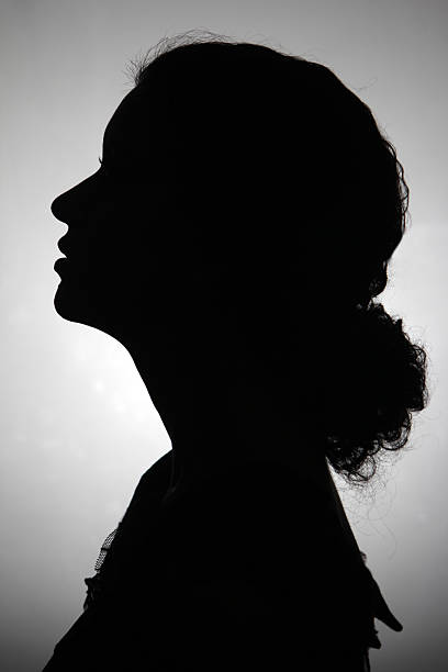 Black and white silhouette of a woman Silhouette of young adult woman.  She is in profile and her long curly hair is tied back.  She is looking slightly up and to the right.  She is against a gray background that is back lit the gray fades to white as it gets closer to the center. woman silhouette stock pictures, royalty-free photos & images