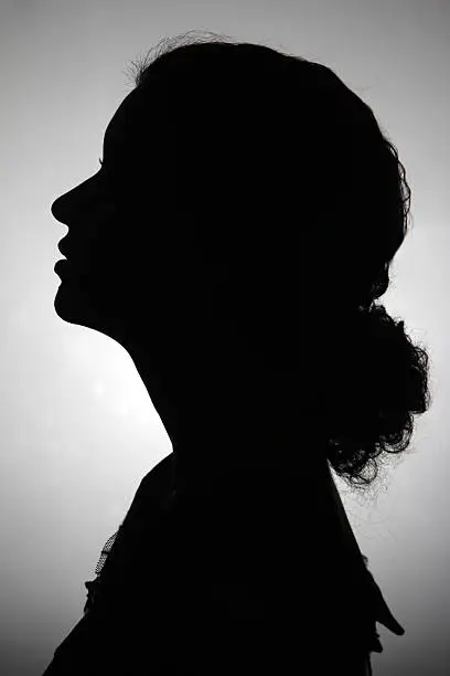 Silhouette of young adult woman.  She is in profile and her long curly hair is tied back.  She is looking slightly up and to the right.  She is against a gray background that is back lit the gray fades to white as it gets closer to the center.