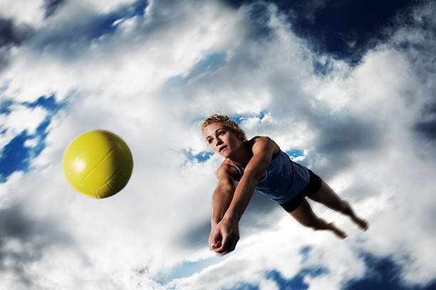 This is a photo of a teenage female athlete diving for a ball on a beach volleyball court in a park setting. This photo is taken from a low angle capturing a great cloudscape behind her. There is some motion blur to this image to add to the feel.