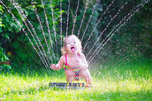 Funny laughing little girl in a colorful swimming suit running though garden sprinkler playing with water splashes having fun in the backyard on a sunny hot summer vacation day