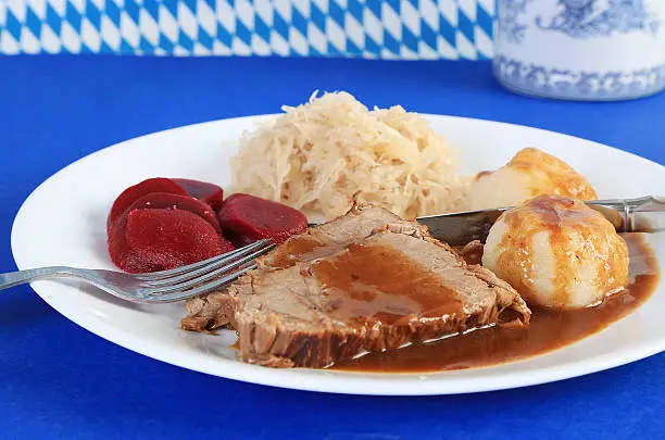 Sauerbraten or German Pot Roast with Potato Dumplings; sauerkraut and pickled beets on blue and white Bavarian colors and beer mug in background
