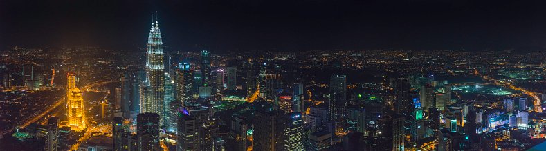 Panoramic view across the futuristic cityscape of Kuala Lumpur, from the iconic twin spires of the Petronas Towers, KLCC park and convention centre to the crowded skyline of hotels, skyscrapers and traffic filled streets illuminated at night, Malaysia. ProPhoto RGB profile for maximum color fidelity and gamut.