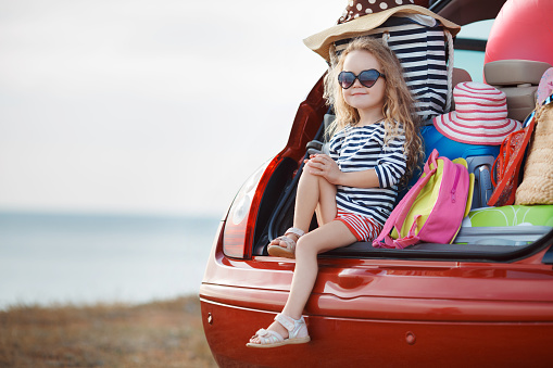 A little girl,a brunette with long curly hair,dressed in a striped sailor shirt,dark sun glasses,and a journey to the sea,sits in the trunk of the red car with clothes,suitcases and bags