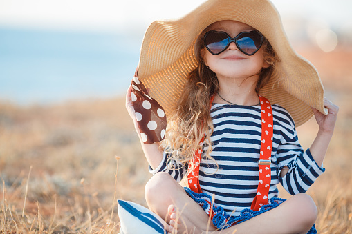 Happy little girl, a brunette with long curly hair, dressed in a striped sailor's t-shirt and red suspenders, wearing dark sun glasses, sitting on a rocky beach in a big straw hat.