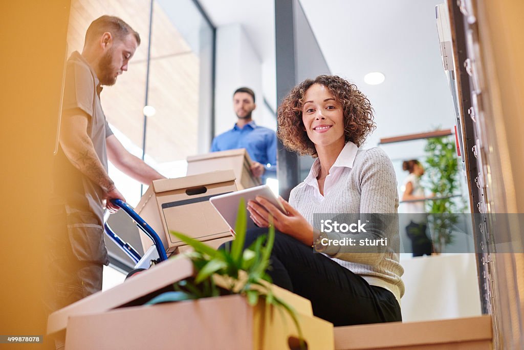 she'll be successful here a mid adult female business owner sits on some moving boxes and looks optimistically at her new offices. Around her , co-workers and removal men are moving boxes and plants as she works on her digital tablet . Change Stock Photo