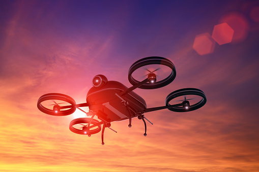 Remote controlled black drone flying in the sky at sunset, with sun behind the silhouette creating a lens flare. The quadricopter hovers mid air thanks to four propellers. The body has a camera, antennas for communication and GPS to follow the route to destination. Blue and orange sky, dusk. Composite image with digitally generated drone.
