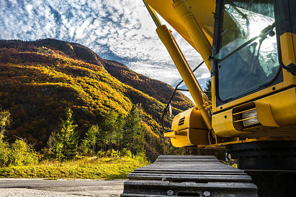 Close-up of a heavy construction machinery (backhoe) in an Alps valley. A large earth moving machine was partially photographed. Visible parts are a cabin, a caterpillar and a hydraulic system. The excavator was photographed in front of a hill overgrown with a dense forests, against a blue sky with scattered clouds at a sunny autumn day.