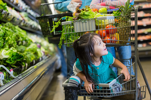 Elementary age Hispanic little girl is smiling while riding in bottom basket of shopping cart in supermarket. Child's parents are pushing shopping cart while shopping for healthy food in grocery store produce section. Cart is filled with fresh fruits and vegetables.