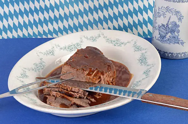 Roast beef in sauerbraten sauce on serving platter with kitchen knife and fork in blue and white colors of South Germany, Bavaria.  Includes beer stein with copy space.