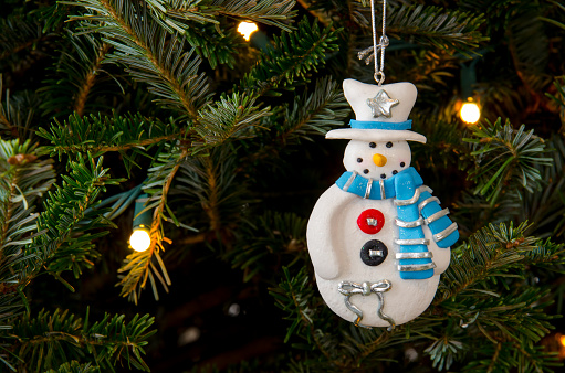 A clay Christmas ornament in the shape of a snowman wears a white hat and blue and silver scarf