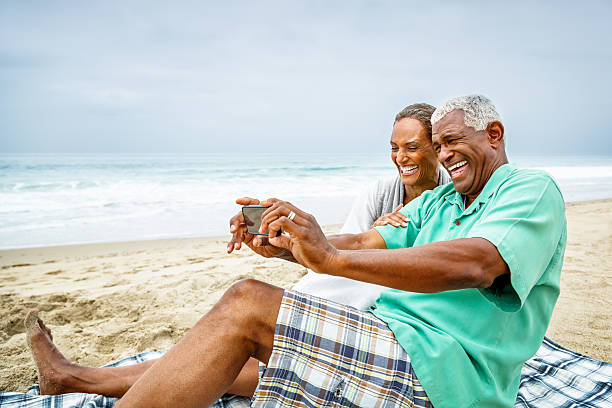 African American couple taking selfie at beach A senior African American couple are sitting on a sandy beach, taking a selfie with a smart phone.  They are both smiling widely as they sit on a plaid blanket over beige sand.  There are white-green sea waters in the background that are topped with a blue-gray sky. lypsela2013 stock pictures, royalty-free photos & images