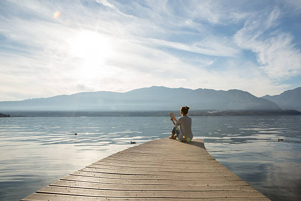 Woman relaxes on lake pier, reads a book Young woman sits on a jetty above the lake, she is reading a book. Beautiful Autumn day in Italy. floating dock stock pictures, royalty-free photos & images