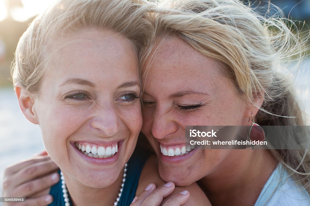 Two women sit on the beach talking and laughing Two friends sit on the beach at sunset talking and laughing. They are close together as the wind blows their hair. Sister Stock Photo