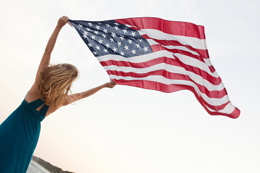 A woman stands with the American flag raised high above her head. The flag is blowing in the wind as she looks off into the distance.