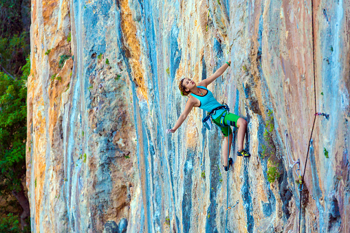 Young Female Climber ascending vertical rocky wall sporty Clothing Blue Shirt Green Pants using Rope and other Safety Gear