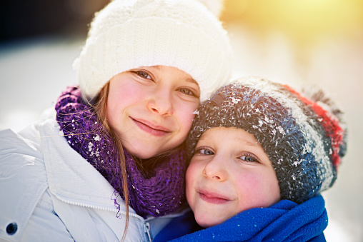 Portrait of brother and sister in winter. Little girl is aged 9 and the boy is aged 5. Kids are embracing, snow visible on the caps.