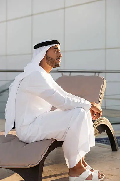 Middle eastern man is sitting on a beach chair on a rooftop of a white building and gazing into the distance, observing the environment and daydreaming. His face and hands are lit by golden sunlight. Image has high contrast. Contains copy space. Made in Dubai, UAE.