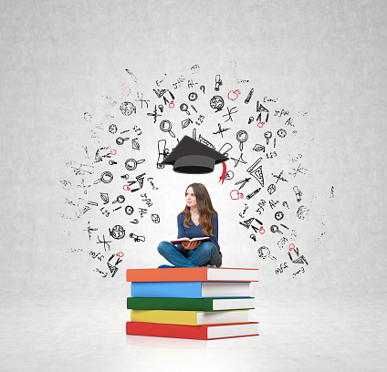 young woman sitting on a pile of books with an open book on her knees thinking about future, square academic hat above her head, white wall with illustrations at the background, concept of education choice