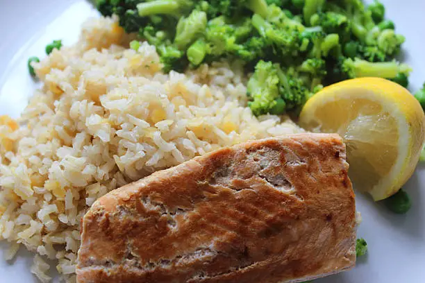 Photo showing a healthy meal consisting of a dry fried salmon fillet served with brown rice and red lentils, garden peas / frozen peas mixed with small pieces of broccoli and a slice of lemon.  This dish is part of a healthy eating, low calorie diet plan.