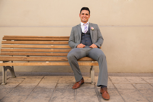 Handsome Hispanic groom sitting on a bench outside and waiting for the bride