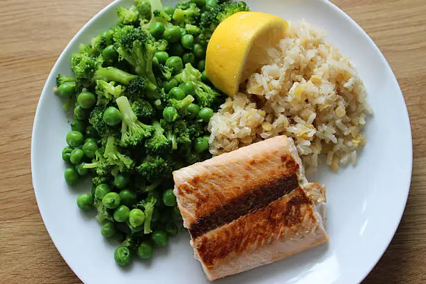 Photo showing a healthy meal consisting of a dry fried salmon fillet served with brown rice and red lentils, garden peas / frozen peas mixed with small pieces of broccoli and a slice of lemon.  This dish is part of a healthy eating, low calorie diet plan.
