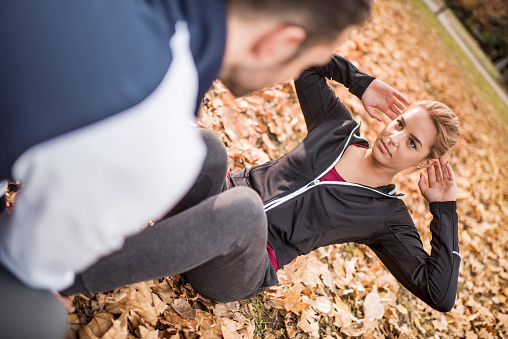 Athletic woman exercising sit-ups with a help of her male friend during autumn day in nature.