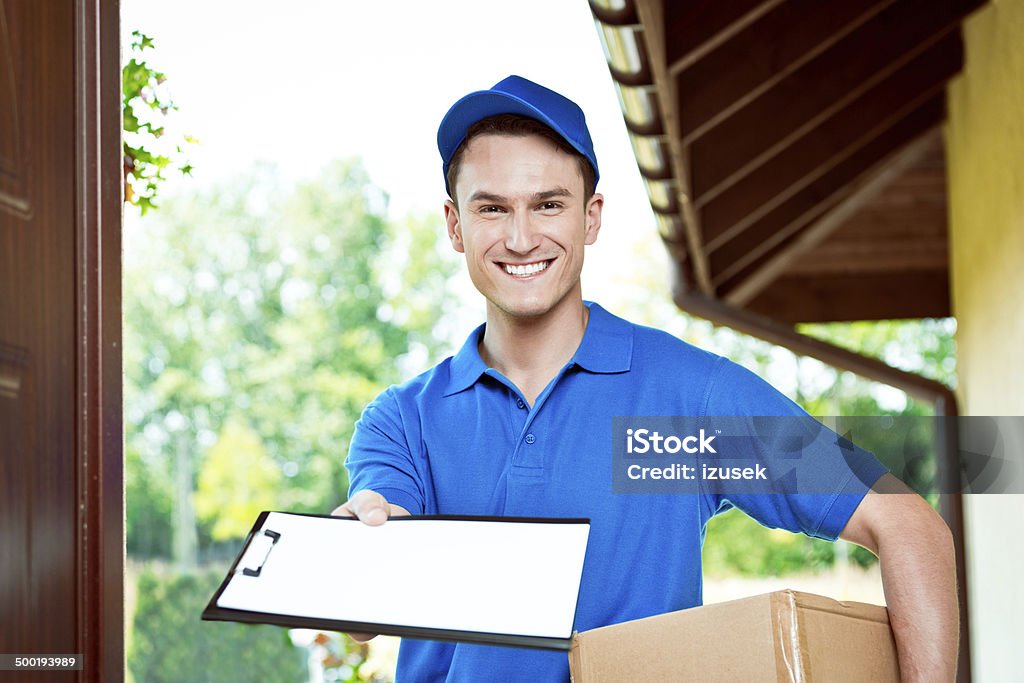 Courier delivering package Delivery man standing at the door of the house and carrying parcel, holding clipboard in hand, smiling at camera. Door Stock Photo