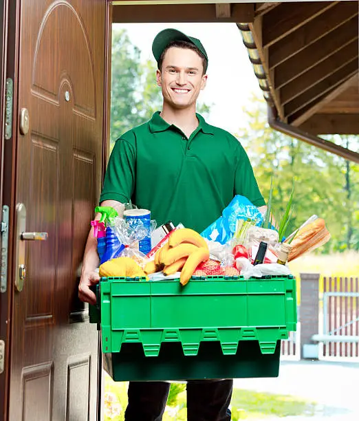 Delivery man standing at the door of the house and carrying box with groceries, smiling at camera.