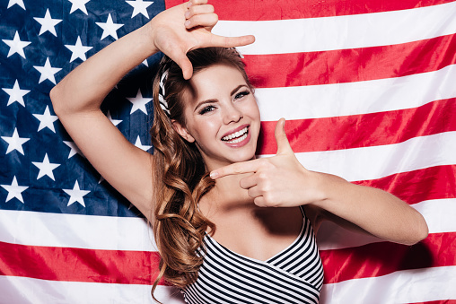 Cheerful young woman making finger frame and looking through it with smile while standing against American national flag